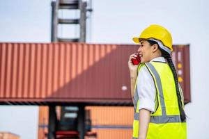 Young confident woman engineer smiling and using radio communication and wearing yellow safety helmet and check for control loading containers box from Cargo freight ship for import and export photo
