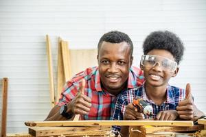 family, carpentry, woodwork, Portrait of cheerful african american father and little son making thumbs up at workshop,  with diverse working tools laying on it photo