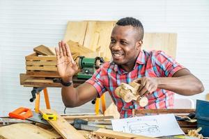 joven carpintero afroamericano sentado en la mesa mostrando modelo de camión de madera y dibujando papel a través de una tableta mientras video en línea chatea con el cliente o enseña en línea en la fábrica