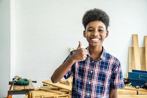 Smiling African-American boy carpenter standing with giving thumbs up as sign of success in a carpentry workshop. photo