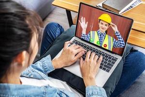 Woman builder in protective helmet waving hand from laptop screen to client. Remote consulting on repair concept while staying at home photo