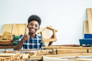 Smiling African-American boy carpenter looks at his own wooden house and thumbs up that helps his father happily do it photo