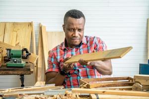 joven carpintero afroamericano mirando y eligiendo madera y usando papel de lija para frotar tablones de madera en la mesa del taller en la fábrica de madera de carpintero foto