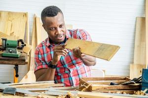 joven carpintero afroamericano mirando y eligiendo madera y usando papel de lija para frotar tablones de madera en la mesa del taller en la fábrica de madera de carpintero foto