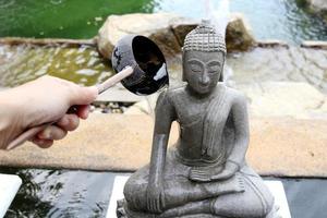 Rock gray color Buddha statue beside pond wet on right side, water flowing from Coconut shell ladle in hand, Thailand.