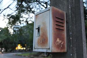 The old metallic box is hanging on cement pole in public park. Outside of box has brown rust. photo