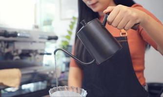 A female cafe operator wearing an apron pours hot water over roasted coffee grounds to prepare coffee for customers in the shop. photo