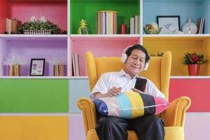 Trendy retired senior man listening music leisurely in smiling face with colorful living room background. Technology elderly lifestyle concept.
