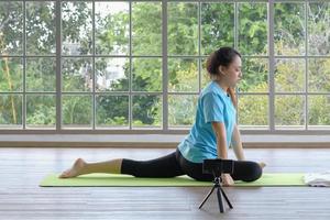 joven asiática concentrada haciendo pose de paloma de yoga siguiendo un tutorial en línea desde un teléfono inteligente en casa o en el fondo del apartamento. enfoque selectivo. foto