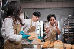 Three young friends and startup partners of bread dough and pastry foods busy with homemade baking jobs while cooking orders online, packing, and delivering on bakery shop, small business entrepreneur