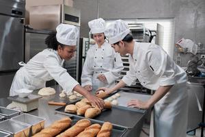 Professional gourmet team, three young chefs in white cook uniforms and aprons knead pastry dough and eggs, prepare bread, and fresh bakery food, baking in oven at stainless steel restaurant kitchen.