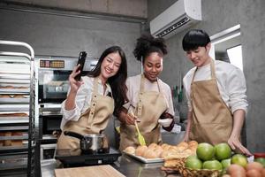 Three young students in cooking class wear aprons enjoyment while taking selfie photo with mobile phone in kitchen, smiling and laughing, preparing eggs and fruits, learn fun culinary course together.