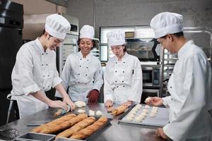 Multiracial professional gourmet team, four chefs in white cook uniforms and aprons knead pastry dough and flour, prepare bread, and bakery food, baking in oven at stainless steel restaurant kitchen. photo