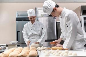 Two professional Asian male chefs in white cook uniforms and aprons are kneading pastry dough and eggs, preparing bread and fresh bakery food, baking in oven at stainless steel kitchen of restaurant. photo