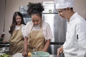 Cuisine course, senior male chef in cook uniform teaches young cooking class students to knead and roll pastry dough, prepare ingredients for bakery foods, fruit pies in stainless steel kitchen. photo