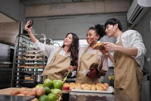 Three young students in cooking class wear aprons enjoyment while taking selfie photo with mobile phone in kitchen, smiling and laughing, preparing eggs and fruits, learn fun culinary course together.