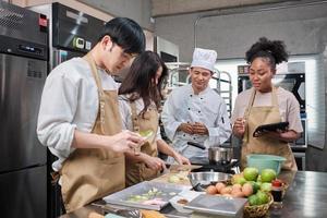 Hobby cuisine course, senior male chef in cook uniform teaches young cooking class students to peel and chop apples, ingredients for pastry foods, fruit pies in restaurant stainless steel kitchen. photo