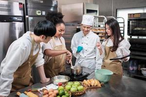 Hobby cuisine course, senior male chef in cook uniform teaches young cooking class students to prepare, mix and stir ingredients for pastry foods, fruit pies in restaurant stainless steel kitchen. photo