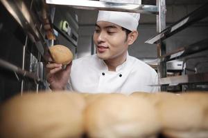 joven chef asiático con uniforme de cocinero blanco y sombrero que muestra una bandeja de pan fresco y sabroso con una sonrisa, mirando su bollo, feliz con sus productos alimenticios horneados, trabajo profesional en la cocina de acero inoxidable. foto