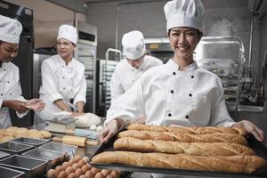 Portrait of young Asian female chef in white cooking uniform looking at camera with a cheerful smile and proud with tray of baguette in kitchen, pastry foods professional and fresh bakery occupation.