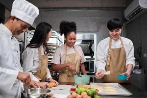 Cuisine course, senior male chef in uniform teaches young cooking class students, brushes pastry dough with eggs cream, prepares ingredients for bakery foods, fruit pies in stainless steel kitchen. photo