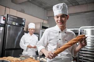 Portrait of young Asian male chef in white cooking uniform looks camera, happy smile and cheerful with baguette, professional foods occupation, commercial pastry culinary jobs in a restaurant kitchen. photo