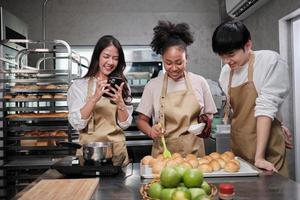 tres jóvenes estudiantes en la clase de cocina usan delantales para disfrutar mientras toman una foto selfie con el teléfono móvil en la cocina, sonríen y ríen, preparan huevos y frutas, aprenden juntos un curso culinario divertido.