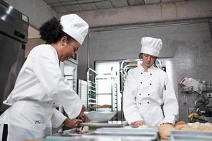 Two professional female chefs in white cook uniforms and aprons knead pastry dough and eggs, prepare bread, cookies, and fresh bakery food, baking in oven at a stainless steel kitchen of a restaurant. photo