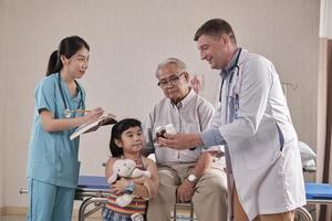 Professional Caucasian male doctor advises grandfather and grandchild, explains prescription with medical trainees at patient hospital bed, illness consultations, and family health examination clinic. photo