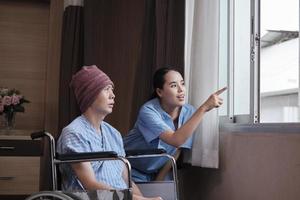 Una joven y uniformada médica de terapia asiática anima a un paciente varón en silla de ruedas en la ventana a apoyar y motivar la recuperación, la enfermedad del cáncer después del tratamiento médico con quimioterapia en la habitación del hospital. foto