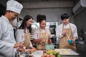 Cuisine course, senior male chef in uniform teaches young cooking class students, brushes pastry dough with eggs cream, prepares ingredients for bakery foods, fruit pies in stainless steel kitchen.