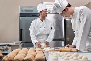 Two professional Asian male chefs in white cook uniforms and aprons are kneading pastry dough and eggs, preparing bread and fresh bakery food, baking in oven at stainless steel kitchen of restaurant. photo