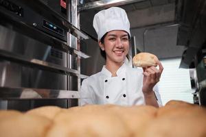 Young Asian female chef in white cook uniform and hat showing tray of fresh tasty bread with smile, looking at camera, happy with his baked food products, professional job at stainless steel kitchen.