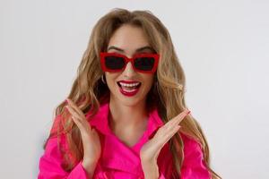 Happy, shock, excited woman face closeup. Girl in bright pink autumn clothes, red sunglasses isolated on white background, copy space. Beautiful female surprised, cheerful. Fun funny model screaming photo