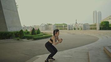 Slim Asian female athlete in black sportswear wearing exercise headphones jumps up stairs in a park near a bridge on a river, urban life.
