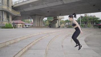 l'atleta femminile asiatica magra in abbigliamento sportivo nero che indossa le cuffie da esercizio salta su per le scale in un parco vicino a un ponte su un fiume, la vita urbana. video