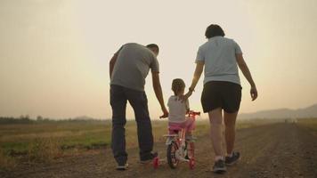 padre y madre asiáticos llevando a su hija en bicicleta rosa en el prado durante las puestas de sol. concepto de familia. video