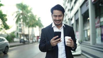 Dieser gutaussehende Mann macht einen langsamen Spaziergang durch die Stadt, lächelt und genießt seinen leckeren Kaffee. tippt Nachrichten auf seinem Smartphone und trinkt heißen Kaffee. trägt einen schwarzen Business-Anzug.