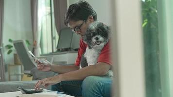 un homme asiatique porte des lunettes, une chemise rouge et un jean avec de l'argent et une calculatrice vérifie les factures, calcule les dépenses, étudie le solde créditeur assis à la table avec un chien à la maison, concept lifestlye finance. video