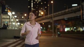 cámara lenta, vista de cámara de seguimiento de fuente. atleta femenina en camisa con capucha trotando en las calles de la ciudad de noche con muchas luces en el fondo. video
