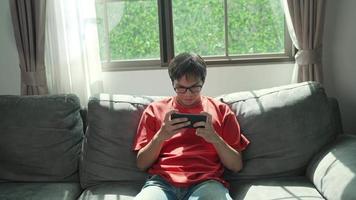 asiatisk man bär rödskjorta glasögon sitter på soffan hemma spelare aktivt spelar mobila games.concept lifestlye teknologi. video