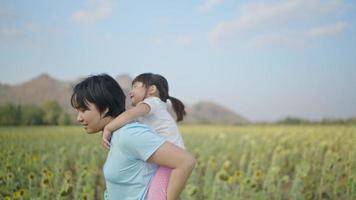 Vidéo 4k au ralenti, une fille asiatique à cheval sur le dos de sa mère a l'air heureuse de marcher dans le champ de tournesols. à l'heure du coucher du soleil. notion de famille. video