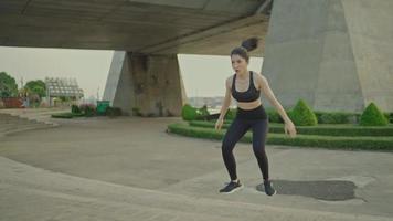 Asian female athlete in black sportswear wearing exercise headphones jumps up stairs in a park near a bridge on a river, urban life.