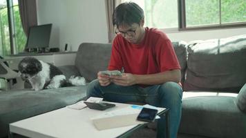 el hombre asiático usa anteojos, camisa roja y jeans con dinero y una calculadora revisa las facturas, calcula los gastos, estudia el saldo de crédito sentado en la mesa con un perro en casa, concetp lifestlye finance. video