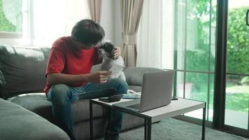 el hombre asiático usa anteojos, camisa roja y jeans con dinero y una calculadora revisa las facturas, calcula los gastos, estudia el saldo de crédito sentado en la mesa con un perro en casa, concetp lifestlye finance. video