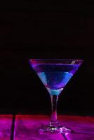 triangular glass with a blue cocktail on a dark background and neon lighting.