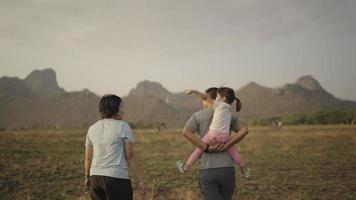 4k-Video in Zeitlupe, Tochter reitet auf dem Rücken des Vaters Mutter steht in der Nähe und kommt glücklich während des Sonnenuntergangs auf der Wiese spazieren. Familienkonzept. video