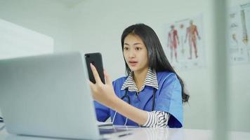 Schöne Ärztin, die an einem Schreibtisch sitzt, mit einem Smartphone Videoanrufe mit Patienten online führt. im Klinikbüro während der Arbeitszeit