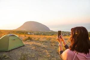 Woman takes photos of the sunrise in the mountains on her phone. Selfie in rising sun. Panoramic view on sea and Ayu-Dag. Camping, outdoor activities, sports mountain hiking, family travel. Crimea.