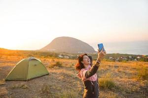 Woman takes photos of the sunrise in the mountains on her phone. Selfie in rising sun. Panoramic view on sea and Ayu-Dag. Camping, outdoor activities, sports mountain hiking, family travel. Crimea.